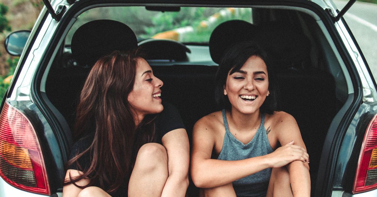 Dwie uśmiechniete młode kobiety siedzące w otwartym bagażniku samochodu (fot. Jorge Saavedra / unsplash.com)