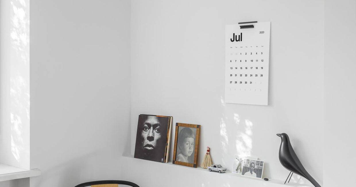 Minimalistyczny kalendarz KAL wiszący na białej ścianie, pod nim półka ze zdjęciami chłopca i albumami winylowymi, widoczne plamy słońca na ścianie
