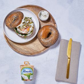 Drewniana taca z talerzem z bajglem z jajkiem sadzonym i serkiem twarogowym Almette z ogórkiem, obok serwetka i nóż, serek Almette