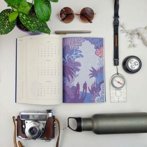 Rozłożony na stole kalendarz krajoznawczy „Na dwór” z ilustracją przedstawiającą parę wędrująca w zimową noc, obok kalendarza leży kompas, bidon, okulary przeciwsłoneczne, aparat fotograficzny