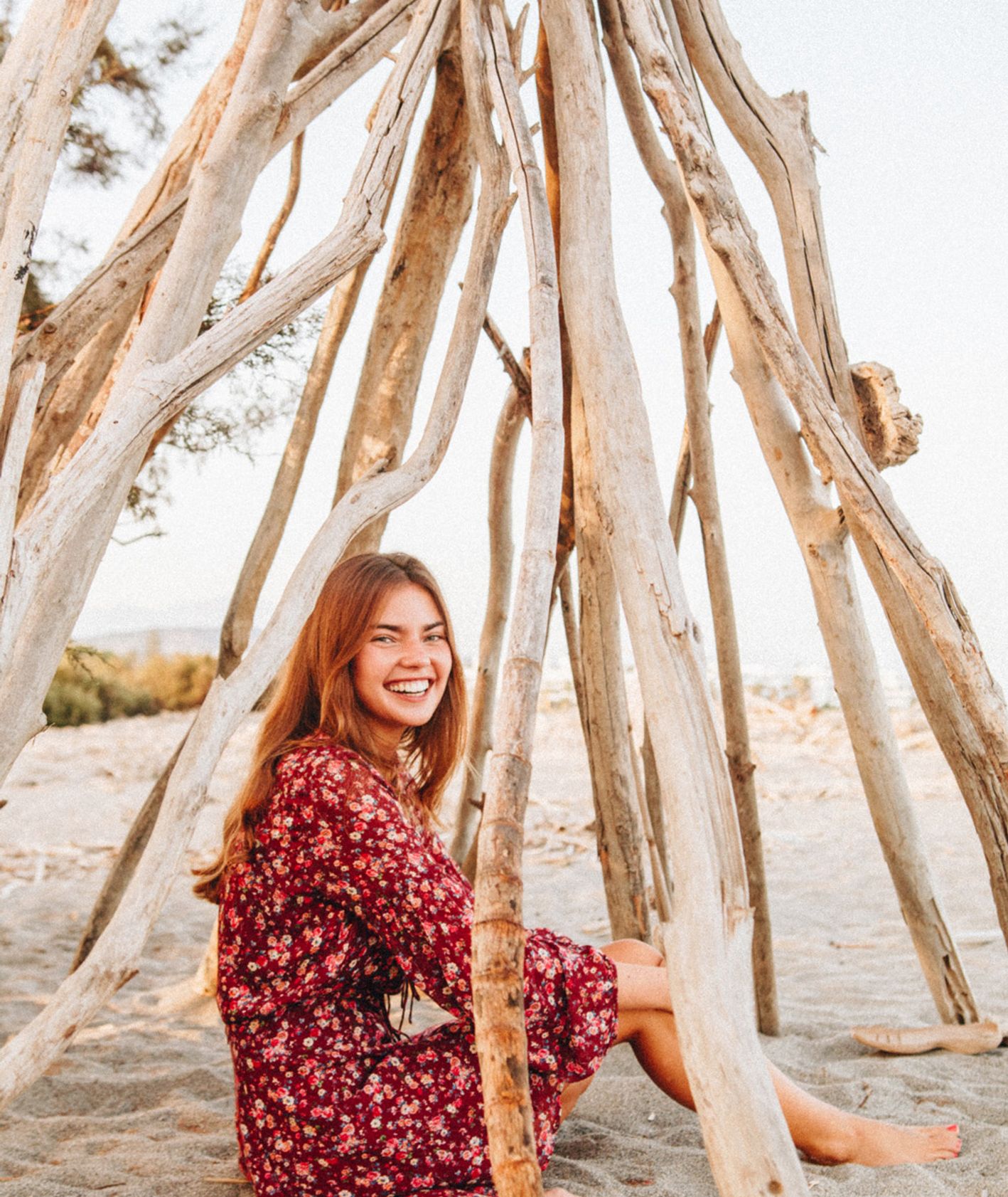 Uśmiechnięta kobieta siedząca pod konstrukcją z gałęzi na plaży. (fot. Sonnie Hiles / unsplash.com)